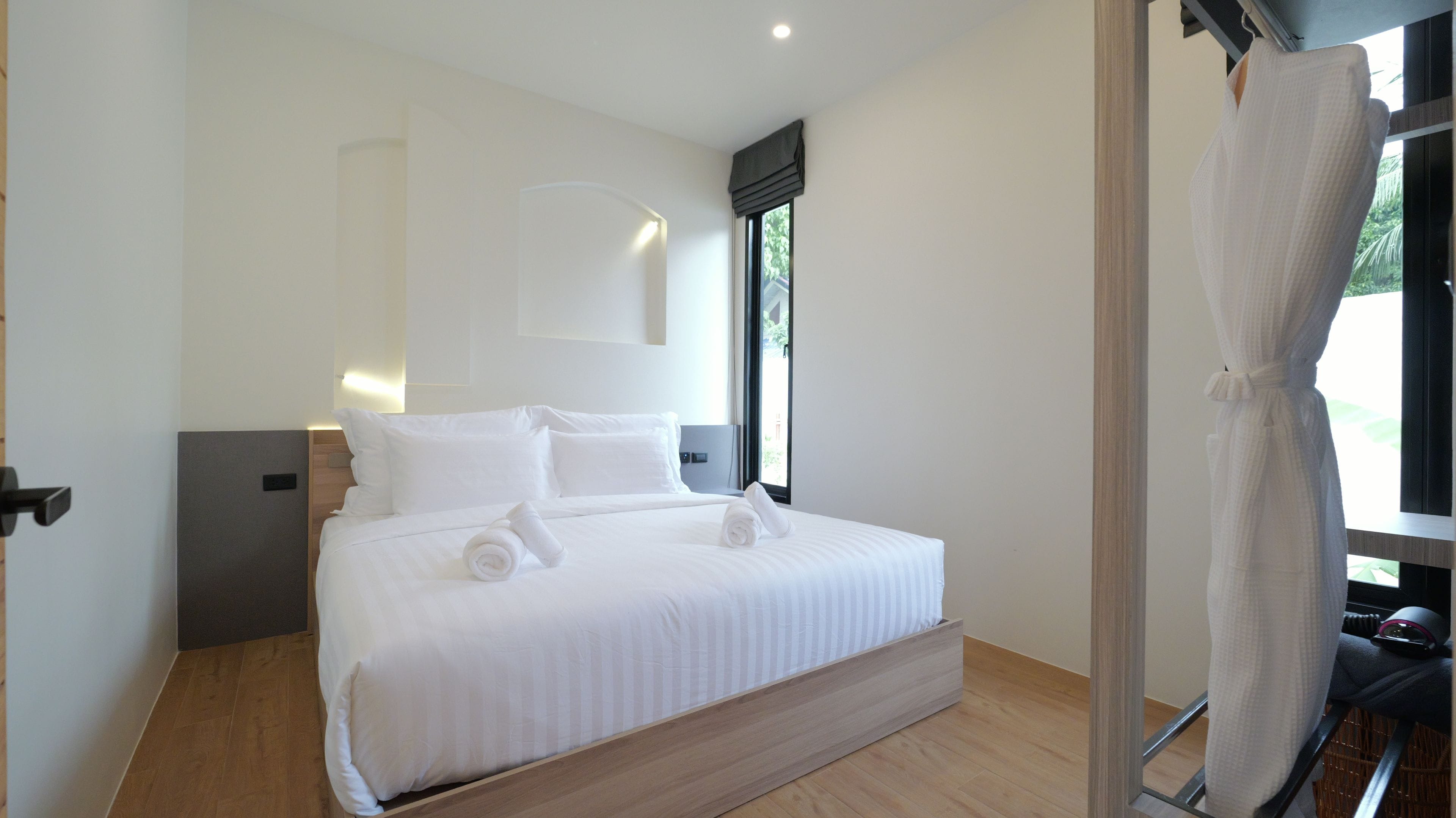 A modern bedroom with a large bed, a bedside table, a wall-mounted light, and a window with a view.