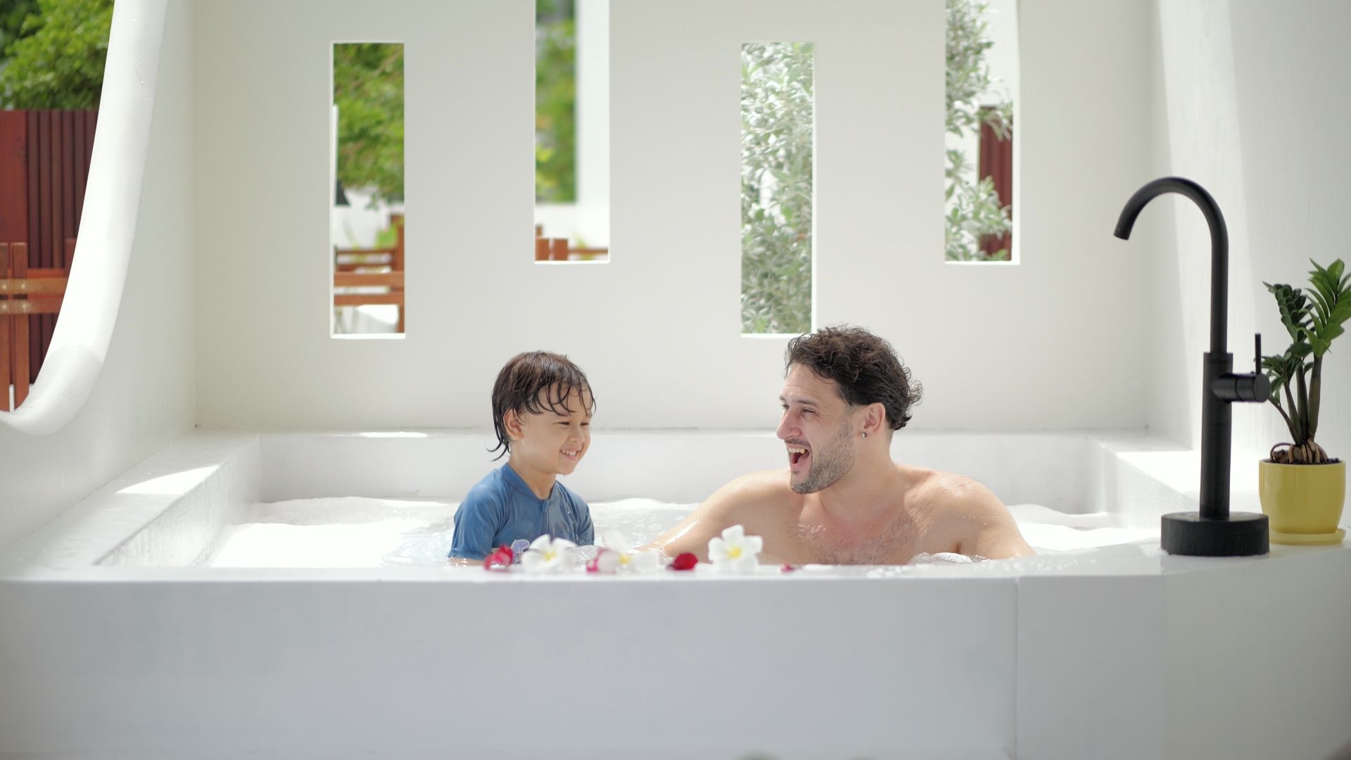 Una bañera con un niño y un adulto adentro, rodeada de ventanas con follaje afuera.