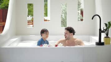 Una bañera con un niño y un adulto adentro, rodeada de ventanas con follaje afuera.