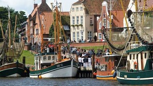 Ein Hafen mit vertäuten Booten, ein Gebäude mit rotem Ziegeldach und Menschen, die sich am Steg versammelt haben.