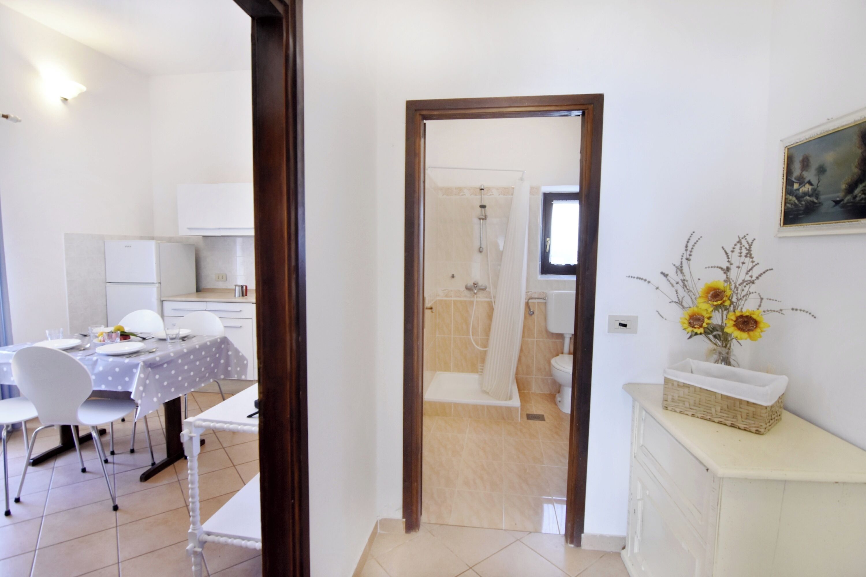 Een kamer met een eettafel, stoelen, een keukenhoekje, een badkamer met douche en een vaas met bloemen op een kast.