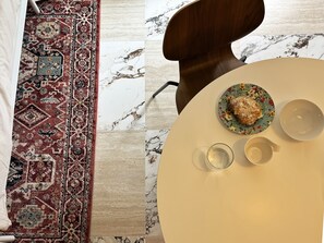 Ein runder Tisch mit einer Tellerplatte mit Blumenmuster, einem Glas und einer Tasse, einem gemusterten Teppich und einem Stuhl mit Holzlehne.