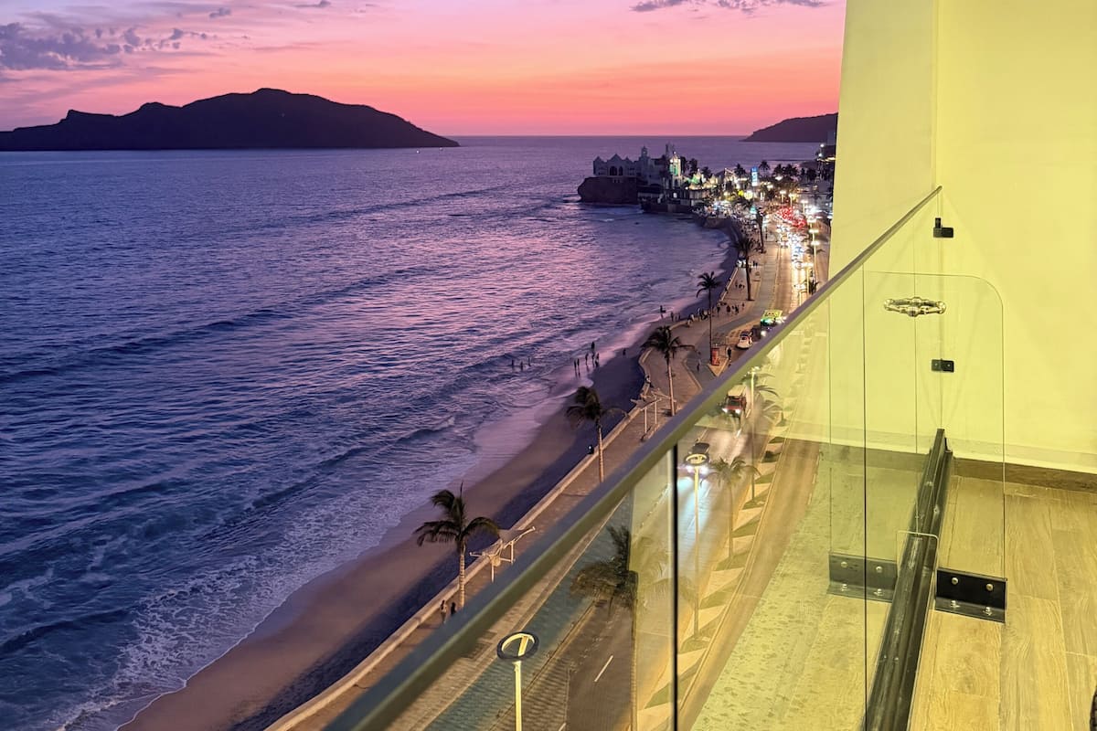 A coastal view from a balcony overlooking a beach with a road parallel to the shore, palm trees, and a distant island.