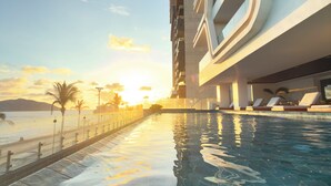 Una piscina con agua cristalina, un edificio moderno con balcones y un atardecer al fondo.