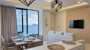 Una sala de estar moderna con un televisor grande, una zona de comedor con una mesa preparada para cuatro personas y un balcón con vistas al mar.