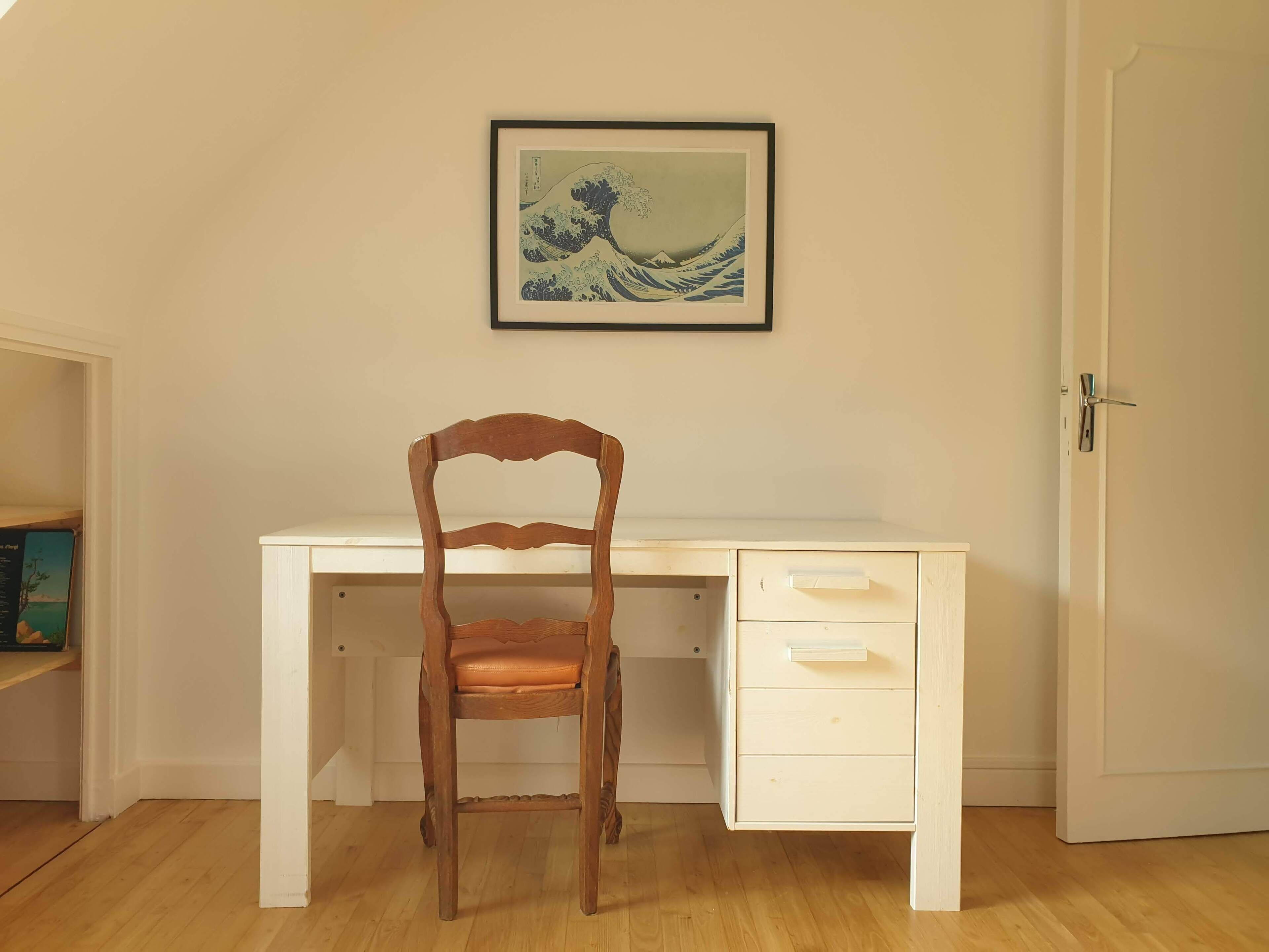A wooden chair in front of a white desk with drawers, a framed artwork above the desk, and a closed door to the right.