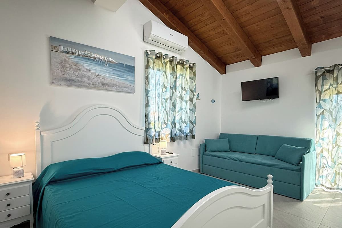 Un dormitorio con cama, mesita de noche, sofá, televisor y un cuadro en la pared.