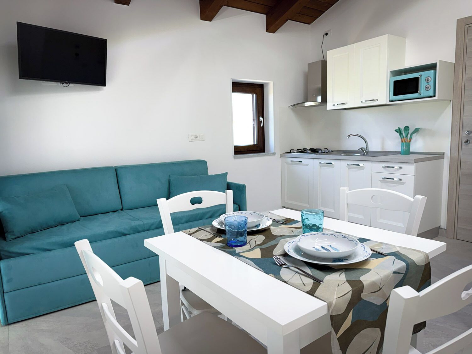 Un espace de vie compact comprenant un canapé de couleur turquoise, une table à manger avec des chaises, un coin cuisine avec un four à micro-ondes et un téléviseur fixé au mur.