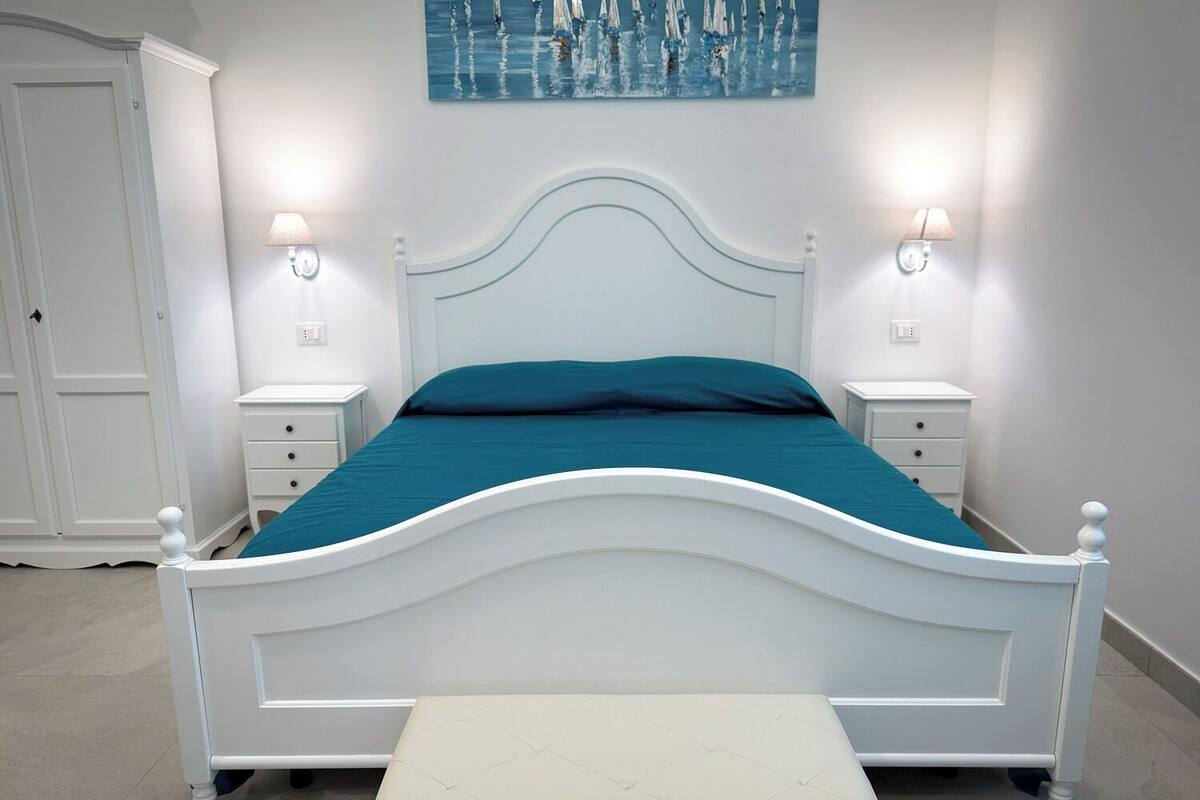Una cama blanca con colcha color turquesa, dos mesitas de noche con lámparas, un banco al pie de la cama y un cuadro en la pared.