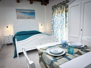 A bedroom with a bed, bedside tables, a table with plates and cutlery, a chair, a window with curtains, and a painting on the wall.