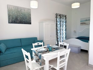 A room with a teal sofa, a white dining table with chairs, a white bed, and a painting on the wall.