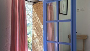 Ein offenes, blau gerahmtes Fenster mit Blick auf Grün, ein Holzstuhl und eine Wandleuchte.