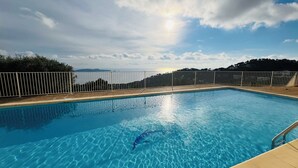 Une piscine à l’eau d’un bleu cristallin, entourée d’une clôture blanche, et offrant une vue sur la mer et les collines en arrière-plan.