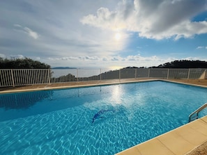 Ein Swimmingpool mit klarem, blauem Wasser, umgeben von einem weißen Zaun, mit Blick auf Meer und Hügel im Hintergrund.