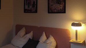 Quarto com cama, almofadas, cobertor, luminária na parede e duas fotos emolduradas.