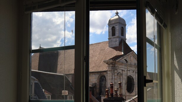 Visão pela janela de um edifício com uma torre de sino, fachada de tijolos e uma pequena janela com uma placa.
