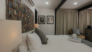 Ein Hotelzimmer mit Bett, Kissen, Kopfteil, einer Wand mit Mosaik, zwei gerahmten Bildern, einer Lampe, einem Sessel und einem Fenster mit Vorhängen.