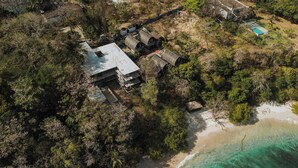 An aerial view of a coastal property with a large house, a swimming pool, and dense surrounding vegetation.