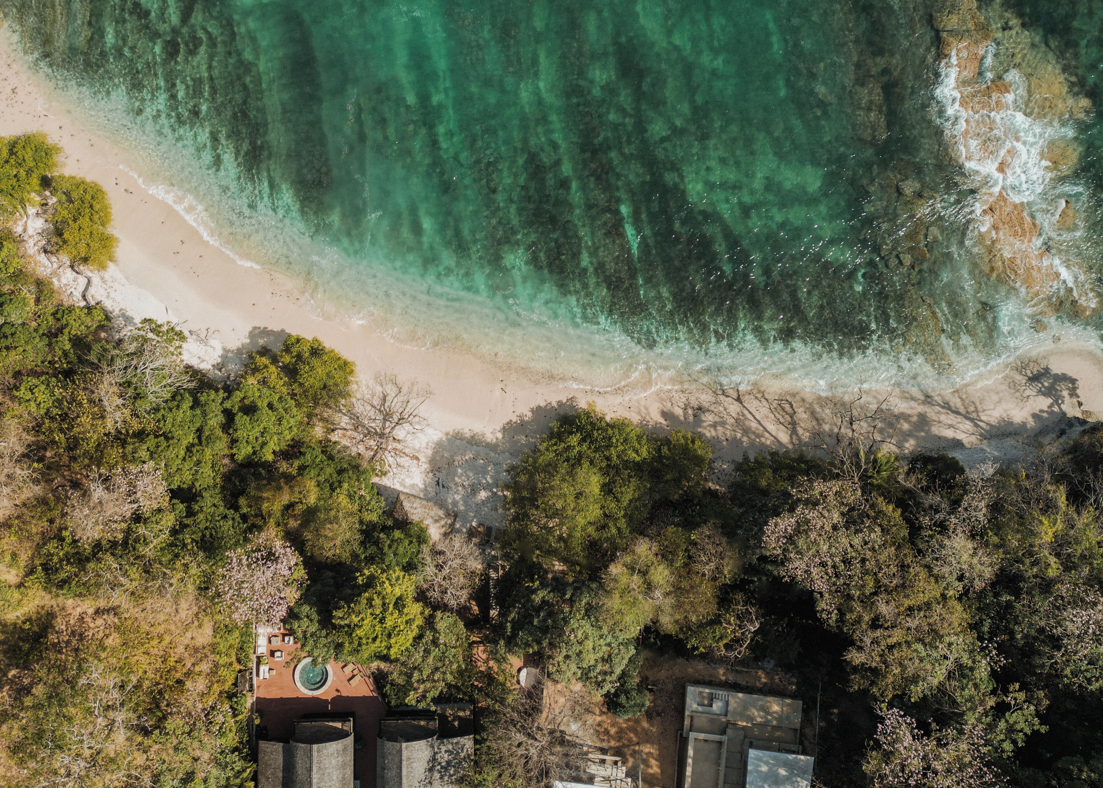 An aerial view of a beach with clear turquoise water, sandy shore, and dense greenery.