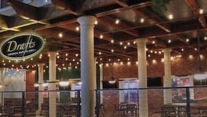 Ulkoterassi, jossa on ruokailualue ja kyltti, jossa lukee ”Drafts Sports Bar & Grill”. Terassilla on tiiliseinä, puupalkit ja valoketju.