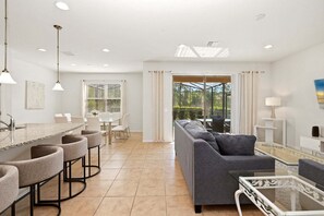 Eine moderne Küche mit Barhocker, ein Essbereich mit Tisch und Stühlen, ein graues Sofa und ein Glastisch.