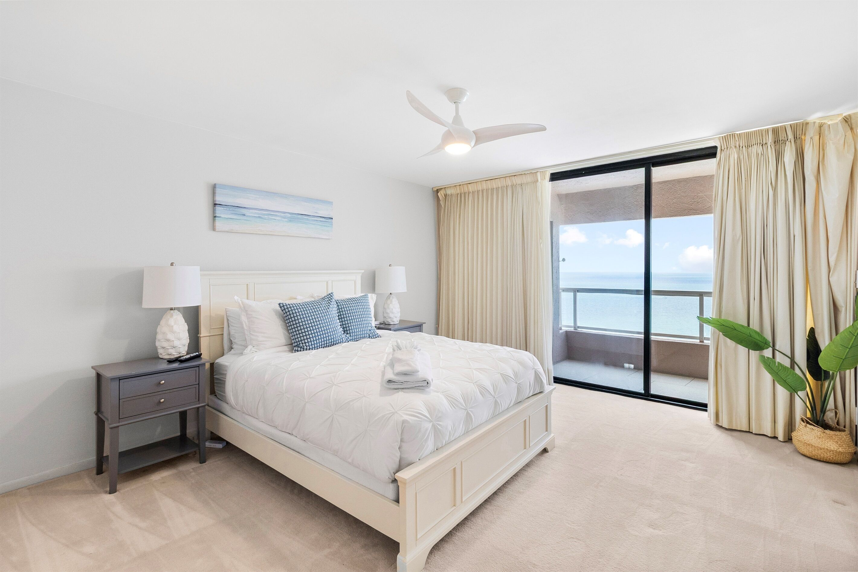A bedroom with a bed, nightstands, a ceiling fan, a view of the ocean, and a balcony with curtains.
