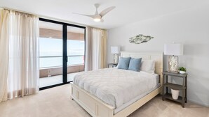 Un dormitorio con cama, mesita de noche, ventilador de techo, ventana con cortinas y vistas al exterior.