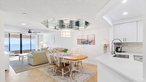 Una cocina moderna con armarios blancos, un comedor con mesa y sillas de madera, un ventilador de techo y vista al océano.