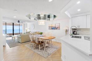 Eine moderne Küche mit weißen Schränken, eine Essecke mit Holztisch und Stühlen, ein Deckenventilator und Meerblick.