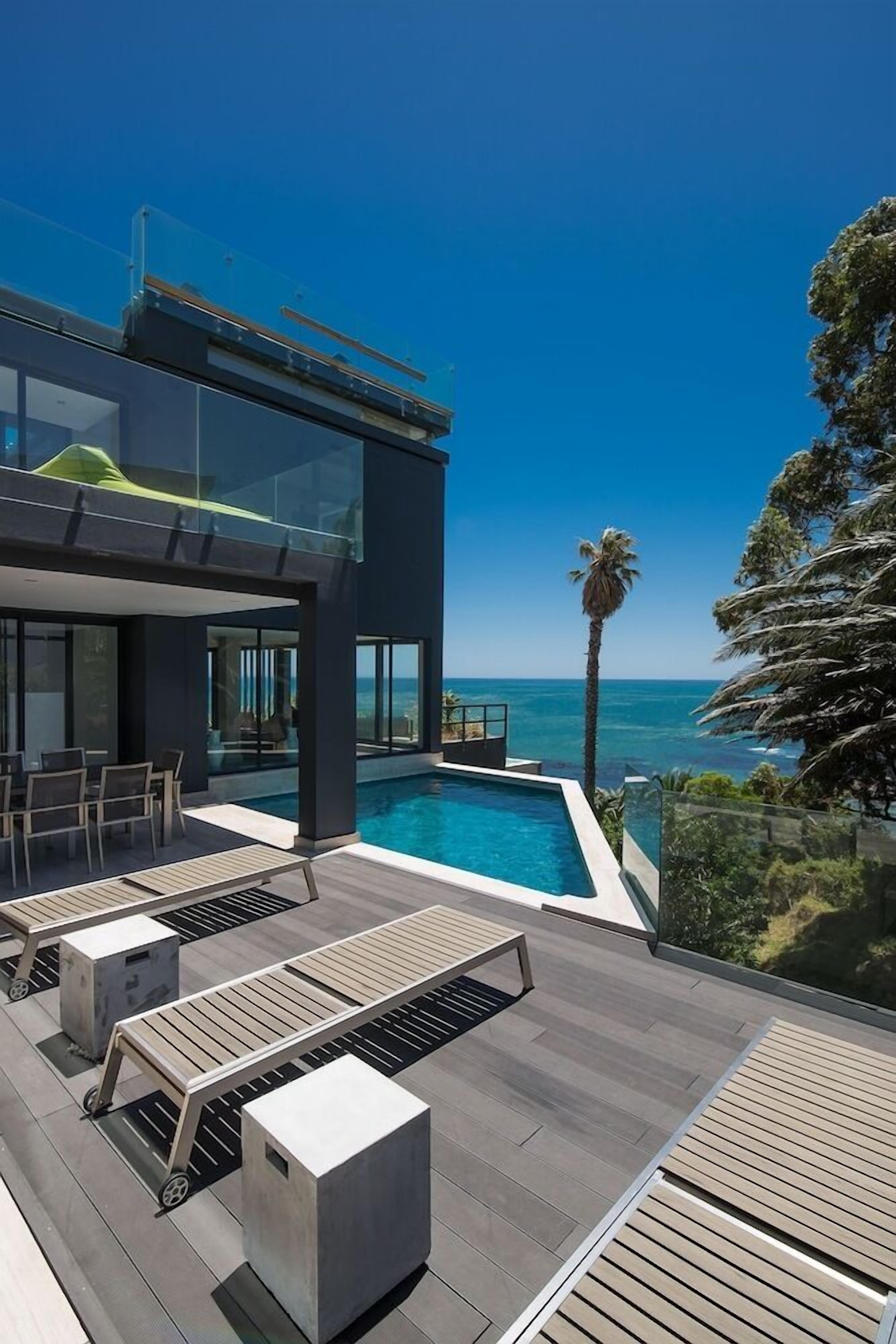 A modern outdoor pool area with a clear view of the ocean, a two-story building, and a palm tree.