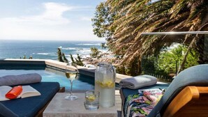 Blick auf einen Pool mit Meerblick, ein Glas und ein Buch auf einer Liege.