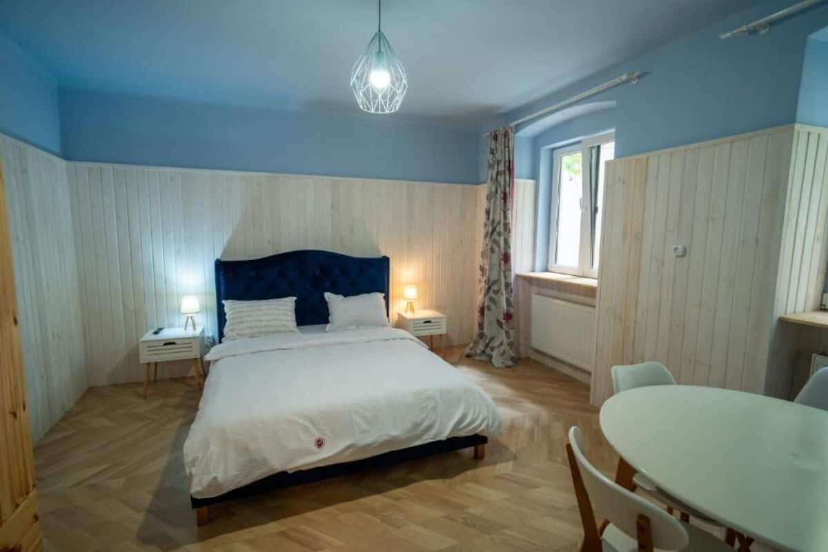 A bedroom with a bed, bedside tables, a hanging light fixture, a window with curtains, and a dining area with a table and chairs.