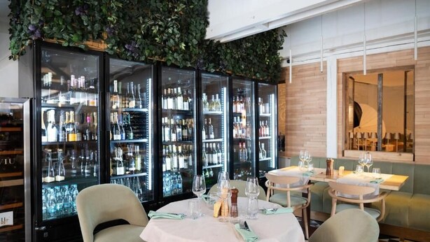 Ein Restaurant mit Barbereich, ausgestattet mit gläsernen Schränken voller Flaschen, einem Essbereich mit runden Tischen für zwei Personen und einer gepolsterten grünen Bank.
