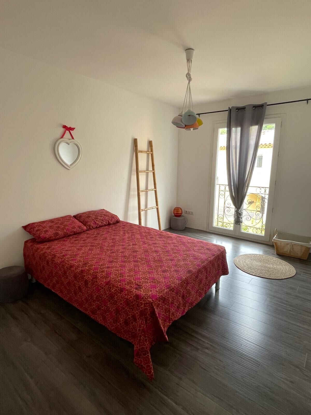 Een slaapkamer met een houten bed, een ladder, een hanglamp, een raam met gordijnen en een klein rond vloerkleed.