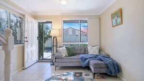 Een woonkamer met een grijze bank, een kamerbreed tapijt en een groot raam met uitzicht op een huis.
