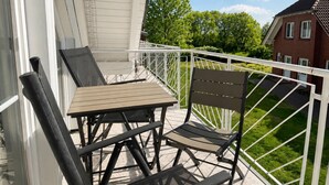 A balcony with a table and two chairs, a white railing, and a view of a brick building and greenery. - Vacation apartment no. 14 in Neddesitz (Neddesitz)