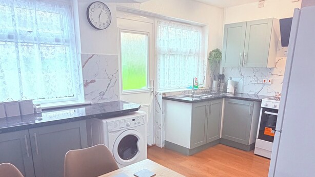 A modern kitchen with a washing machine, a dining table with chairs, and a clock on the wall. - House In Cannock-3 Bed Sleeps 5 (Cannock)