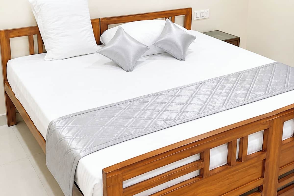A wooden bed with a white mattress and gray pillows, a quilted blanket, and a bedside table with a lamp.
