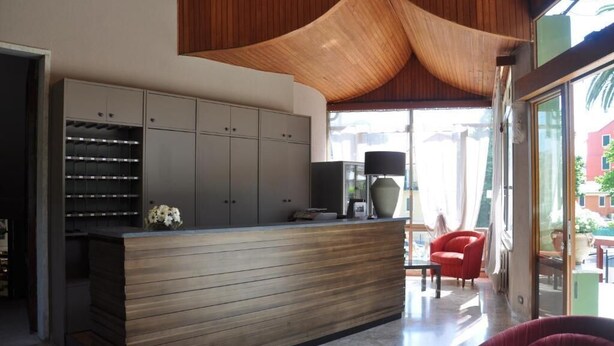 A modern reception area with a wooden reception desk, grey cabinets, and a large window offering a view of the outside. - Hotel Residence Moneglia (Moneglia)