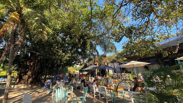 Un área de comedor al aire libre con sillas y mesas de madera, rodeada de árboles.