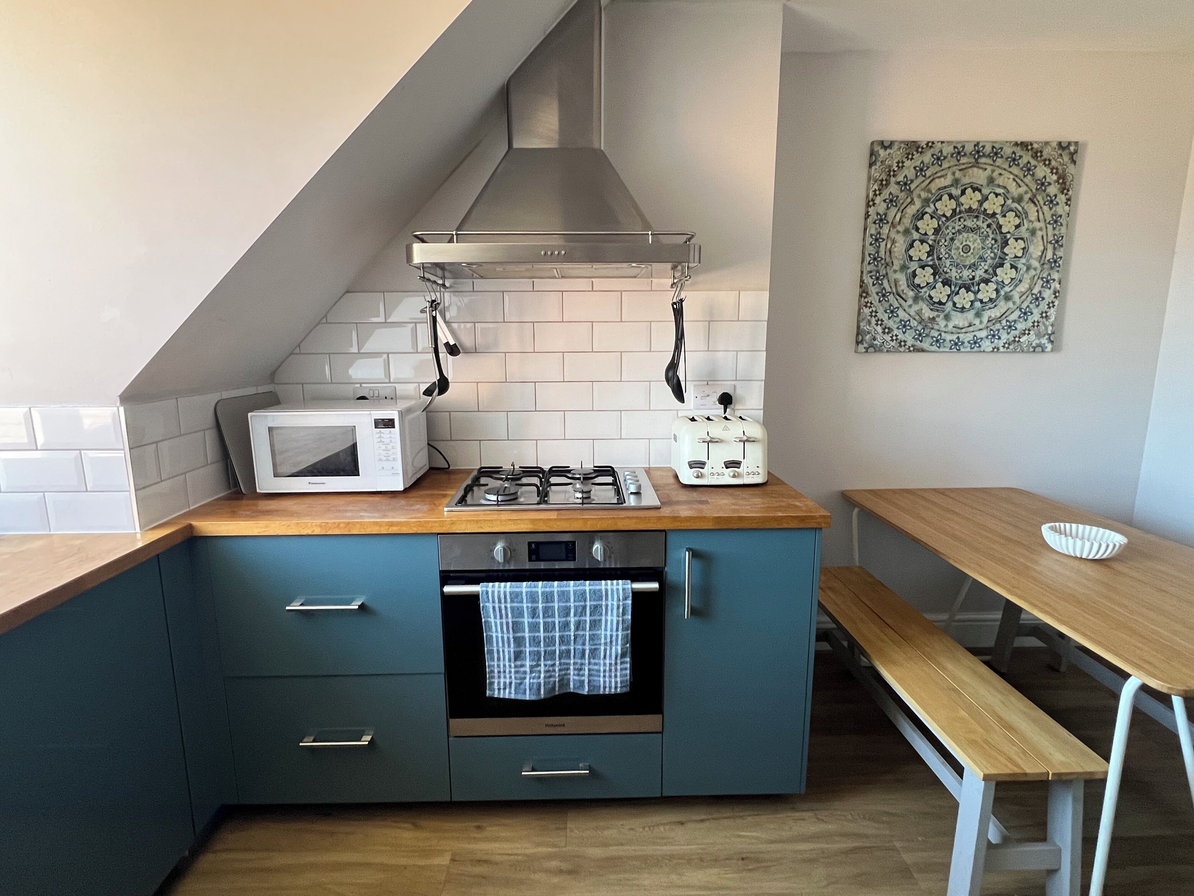 A compact kitchen with a microwave, stove, and toaster on a wooden countertop, a bench, and a patterned wall hanging.