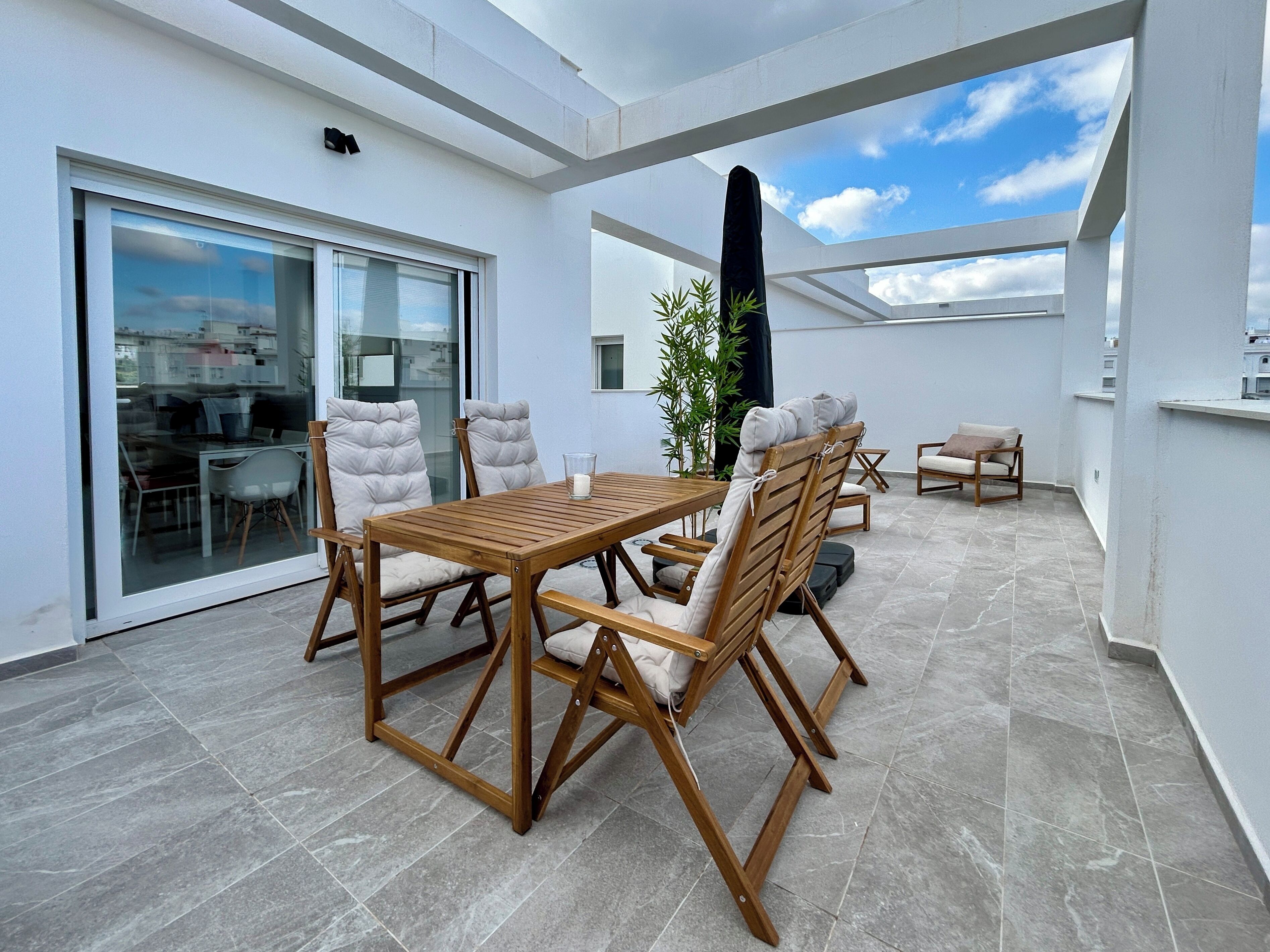 A modern outdoor patio with a wooden table and chairs, a glass door leading to an interior space, and a view of the sky.