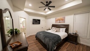Une chambre à coucher comprenant un lit, un ventilateur de plafond, une commode, un miroir, un téléviseur et une fenêtre avec des stores.