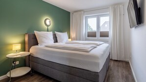 Ein ordentlich bezogenes Bett in einem Zimmer mit grüner Wand, einem kleinen Nachttisch mit Lampe und einem Fenster mit Vorhängen.