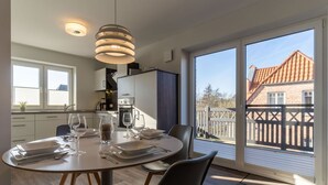 Ein Essbereich mit Tisch für zwei Personen, eine Küche im Hintergrund und Blick auf ein Haus durch große Fenster.