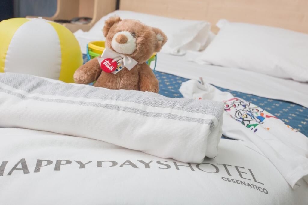 A bed with a white sheet labeled 'HAPPY DAYSHOTEL CESENATICO,' a brown teddy bear with a tag, and a yellow and white striped beach ball.