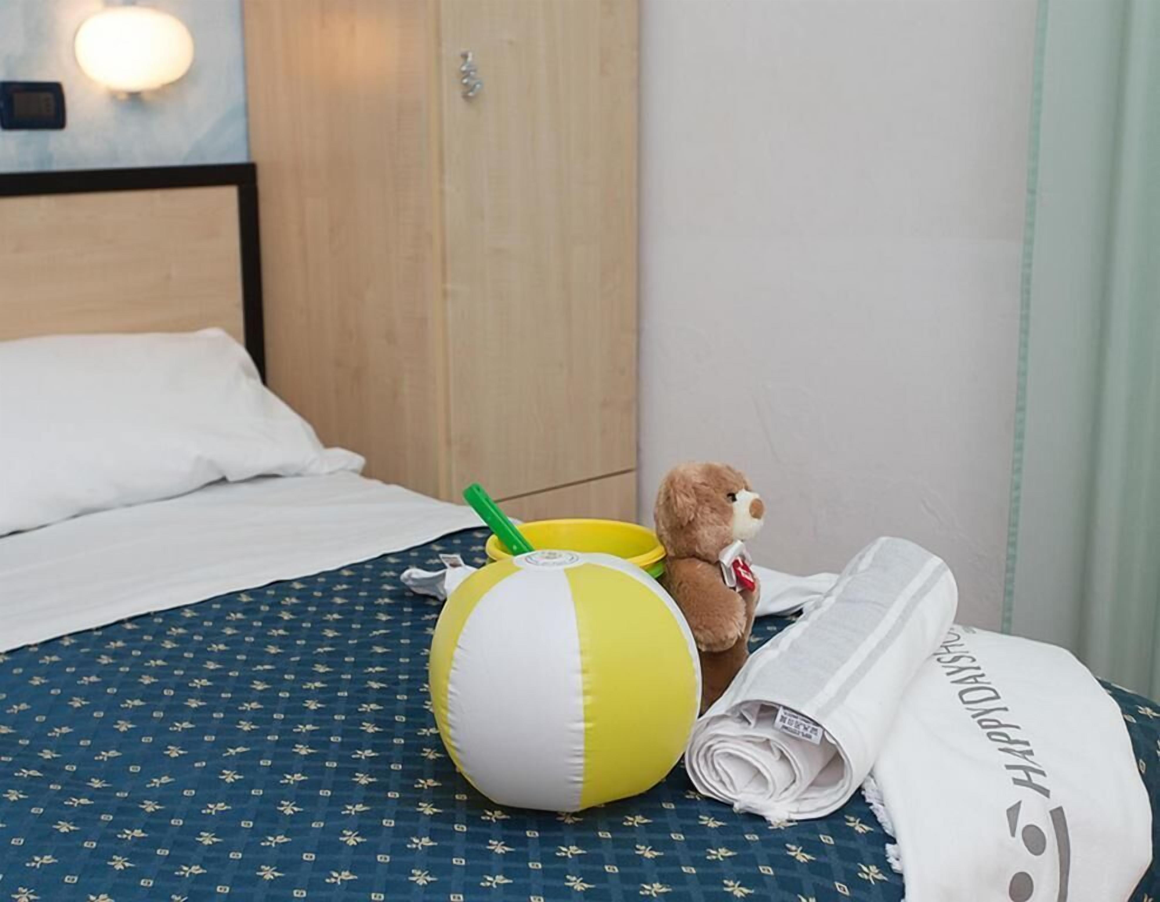 A bed with a blue patterned bedspread, a yellow and white beach ball, a teddy bear, and a rolled-up newspaper on the bed.