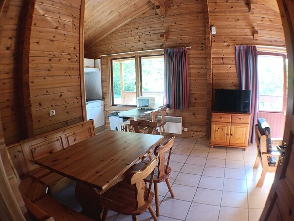 Interior de una cabaña de madera con una mesa de comedor, sillas, una zona de cocina y un televisor.