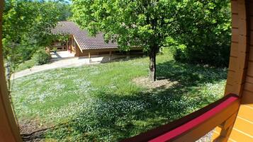 Une vue à travers une fenêtre d’une structure en bois, avec un toit recouvert de tuiles, un arbre et une zone herbeuse parsemée de fleurs blanches.