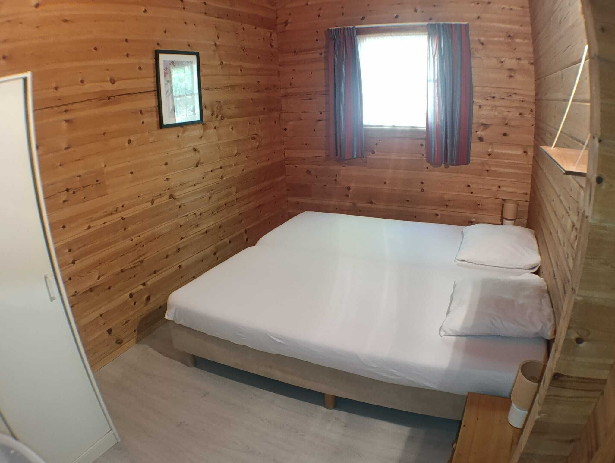 Habitación tipo cabaña de madera con cama, ventana con cortinas e imagen enmarcada en la pared.